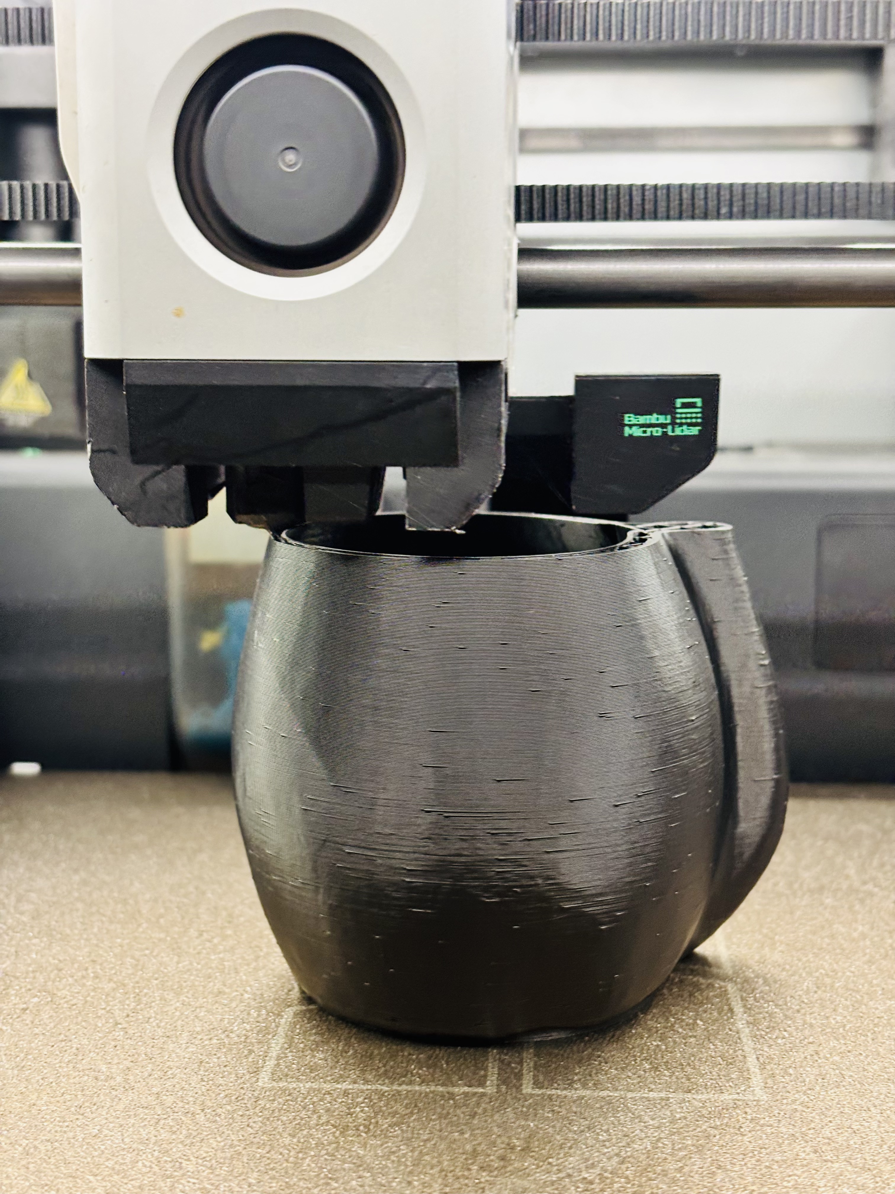 Nahaufnahme eines Bambu Lab-Druckkopfes, der Schichten auf ein abgerundetes, schwarzes 3D-gedrucktes Objekt aufträgt.