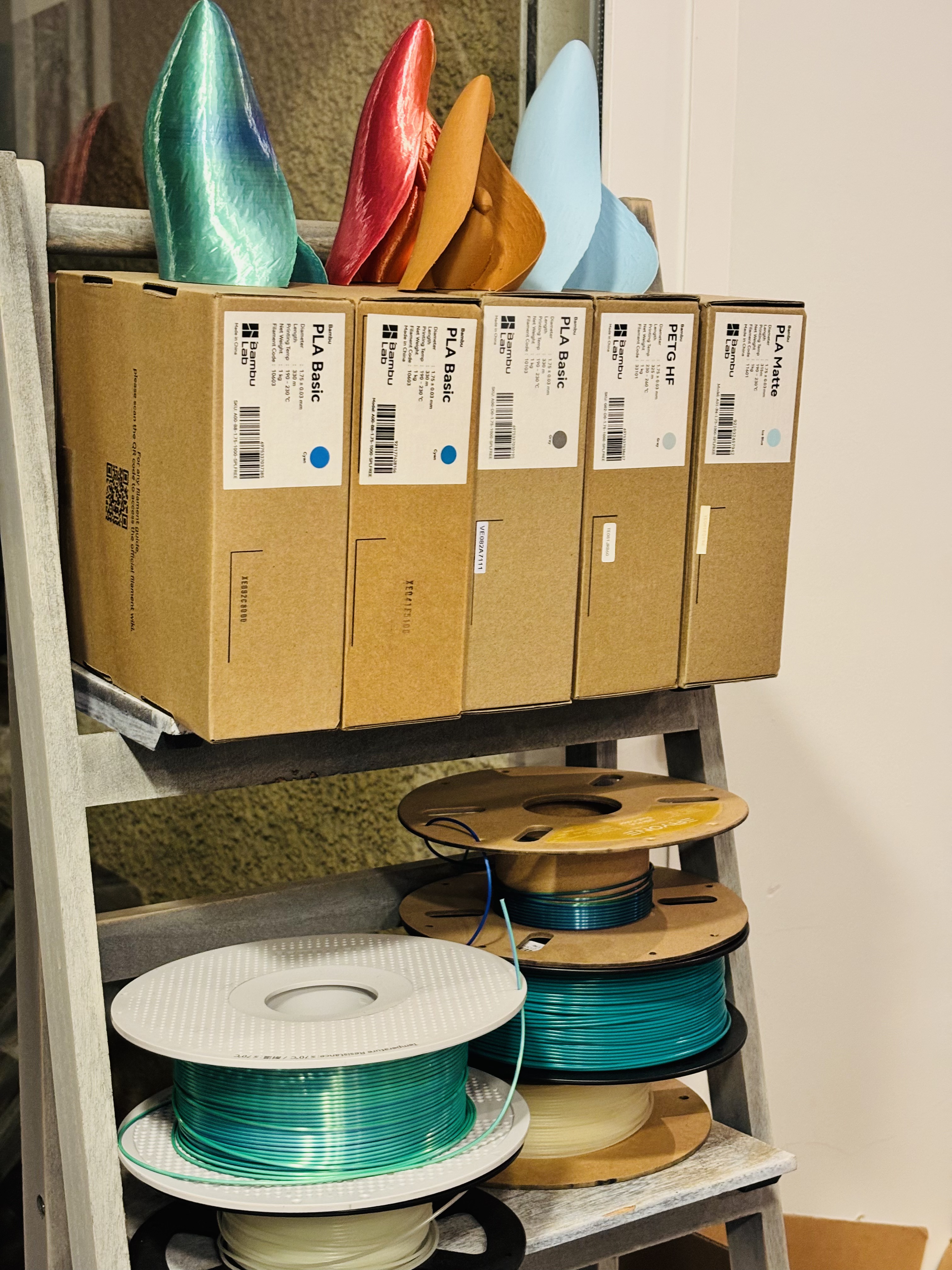 Leiterregal mit farbenfrohen, 3D-gedruckten Ausstellungsstücken oben und Schachteln und Spulen mit Bambu Lab PLA-Filament unten
